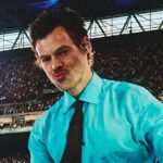 Harry Styles Earns Guinness World Record with 12 Sold-Out Wembley Shows Male performer in a turquoise shirt and black tie taking a selfie on stage with a large crowd behind him in a stadium.