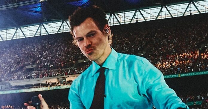 Harry-Style Male performer in a turquoise shirt and black tie taking a selfie on stage with a large crowd behind him in a stadium.