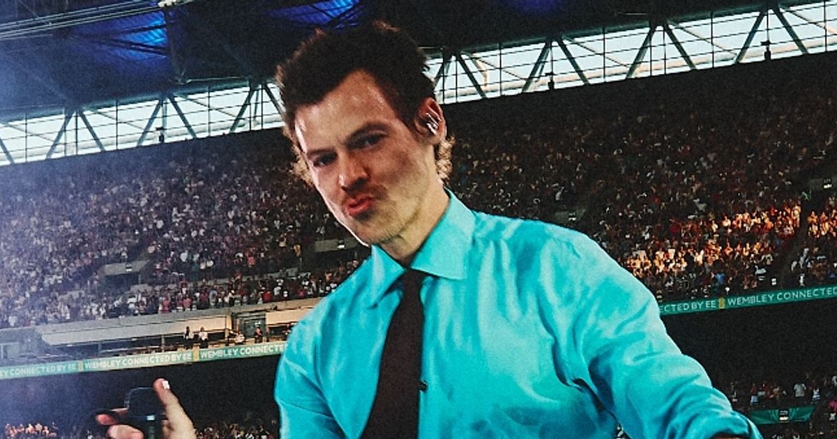 Harry-Style - THEWILL NEWS MEDIA Male performer in a turquoise shirt and black tie taking a selfie on stage with a large crowd behind him in a stadium.