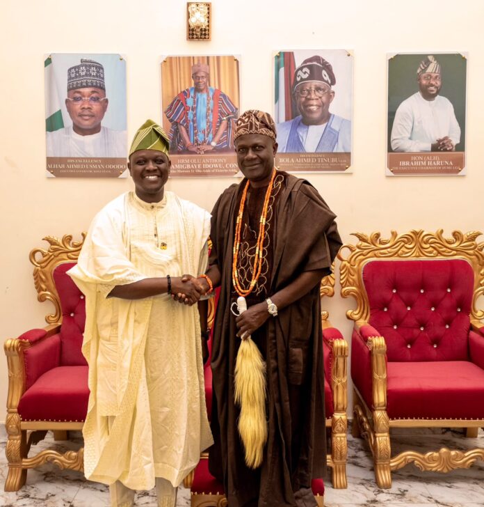 1002761345 Two men in traditional Nigerian attire shake hands in a formal room with gold-trimmed red chairs and portraits on the wall behind them.