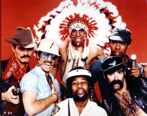 170819_village_people Six men in colorful 70s‑80s costumes posing together against a red background, including a cowboy, construction worker, Native American chief, biker, and sailor.