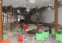 Four Killed, 10 Injured In Explosion At Damascus Café Interior of a ruined cafeteria with scattered debris, broken ceiling tiles, and toppled chairs in bright green, orange, and red; damaged walls and fallen fixtures indicate a severe collapse event.