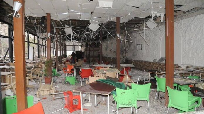 Interior of a ruined cafeteria with scattered debris, broken ceiling tiles, and toppled chairs in bright green, orange, and red; damaged walls and fallen fixtures indicate a severe collapse event.