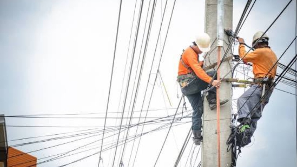 Two utility workers wearing hard hats climb a tall concrete pole amid a tangle of power lines.