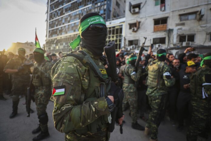 Masked soldier in camouflage with a green headband and rifle in a crowded urban street.