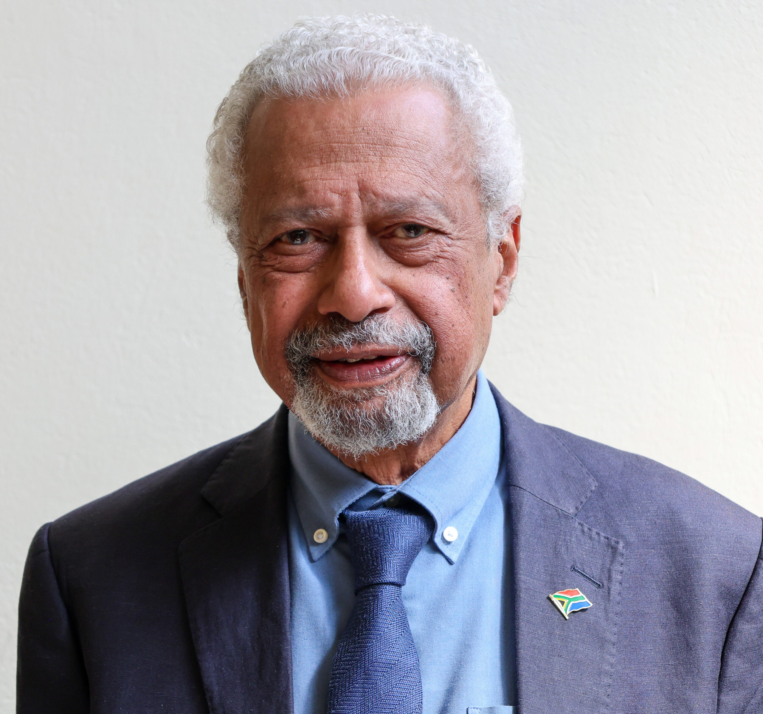 Close-up portrait of an older man with white hair and beard, wearing a dark blazer, blue shirt, and tie, with a South Africa flag lapel pin visible on the lapel.