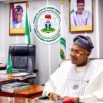 Businessman Alleges N400m Scam By Self-Styled PFIPC DG as Reps Deepen Probe Man in traditional Nigerian attire sits at a desk in a government office with flags and portraits in the background, looking to the side.