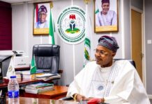 Businessman Alleges N400m Scam By Self-Styled PFIPC DG as Reps Deepen Probe Man in traditional Nigerian attire sits at a desk in a government office with flags and portraits in the background, looking to the side.