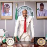 OPINION: PFIPC Scandal and the Urgent Reforms Required Man in a white suit and red tie leans on a polished desk in a formal office, with Nigerian flags and framed portraits behind him.