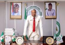 OPINION: PFIPC Scandal and the Urgent Reforms Required Man in a white suit and red tie leans on a polished desk in a formal office, with Nigerian flags and framed portraits behind him.