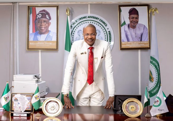 Man in a white suit and red tie leans on a polished desk in a formal office, with Nigerian flags and framed portraits behind him.