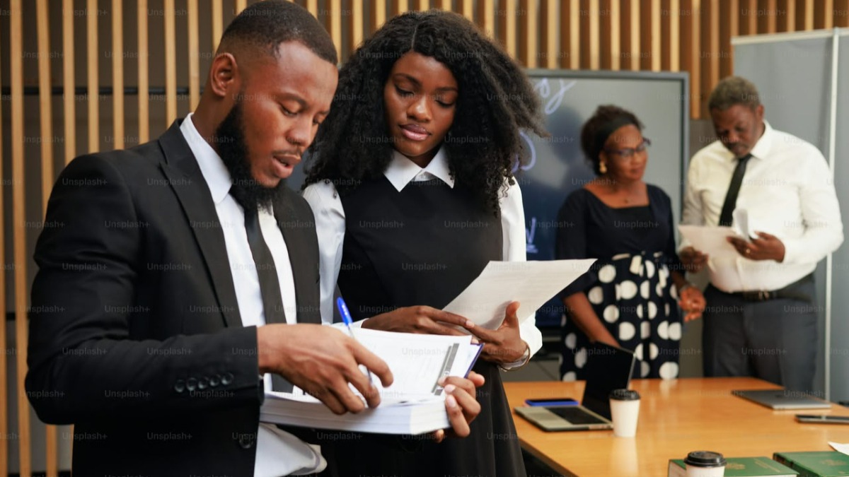Two professionals in business attire review and sign documents at a table in a conference room.