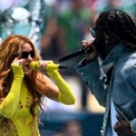 Burna Boy Hits New Career High As First African Artist To Top Billboard Global 200 Two performers sing into wireless mics on an outdoor stage; woman in neon yellow cutout bodysuit beside a man with dreadlocks.