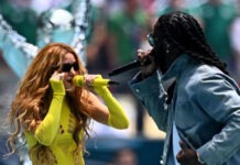 Burna Boy Hits New Career High As First African Artist To Top Billboard Global 200 Two performers sing into wireless mics on an outdoor stage; woman in neon yellow cutout bodysuit beside a man with dreadlocks.