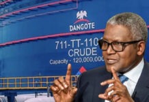 Dangote Refinery Completes $2.5bn Private Placement Ahead Of Planned IPO Man in a dark suit and glasses making a two-handed gesture while speaking, with a large blue Dangote crude oil tank in the background.