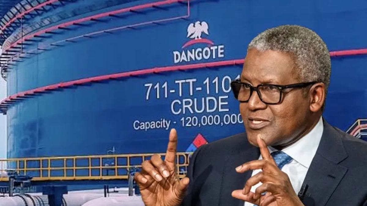 Man in a dark suit and glasses making a two-handed gesture while speaking, with a large blue Dangote crude oil tank in the background.
