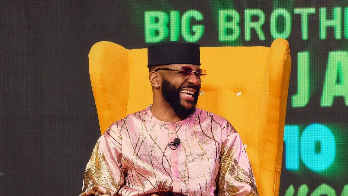 _Ebuka Obi Uchendu - THEWILL NEWS MEDIA Bearded man in a pink traditional outfit and black cap laughing while seated in a bright yellow high-back chair on a Big Brother Naija stage, mic clipped to his chest.