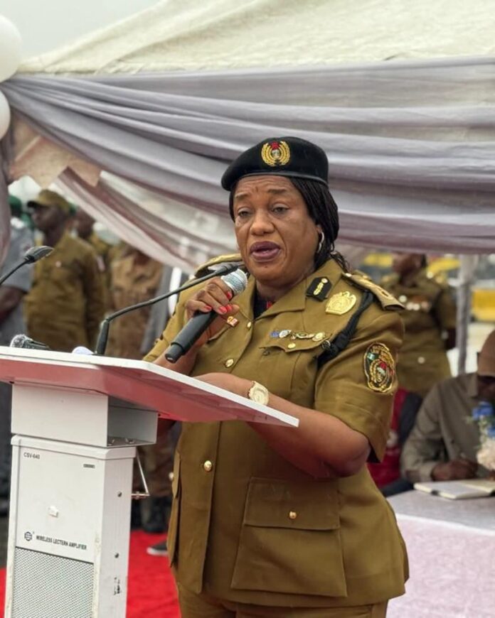 Female military officer in khaki uniform delivers a speech at a podium, holding a microphone, with soldiers in the background under a fabric canopy.