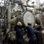 Nigeria’s Crude Oil Output Reaches Highest Level Since 2020 as Questions Mount Over Economic Impact Two workers in hard hats inspect a large valve amid a dense network of industrial pipes and equipment at an oil/refinery facility.