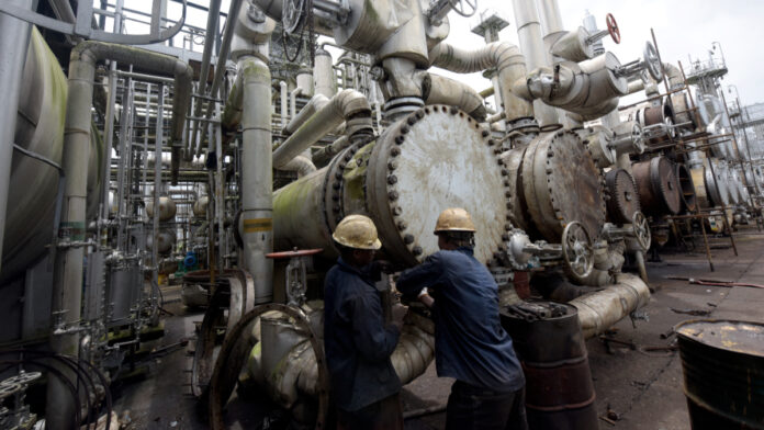 Two workers in hard hats inspect a large valve amid a dense network of industrial pipes and equipment at an oil/refinery facility.