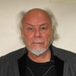 Ex-UK Pop Star Gary Glitter Charged With Historical Sexual Offences Dating Back to 1978 Older man with white goatee and balding head, wearing a gray blazer over a black shirt, posing against a light wall.