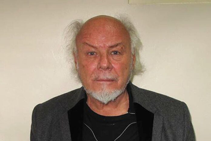 Gary Glitter Older man with white goatee and balding head, wearing a gray blazer over a black shirt, posing against a light wall.