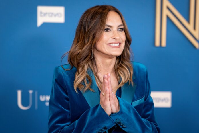 Mariska Hargitay Smiling woman in a blue velvet blazer with hands pressed together in a greeting against a blue event backdrop.