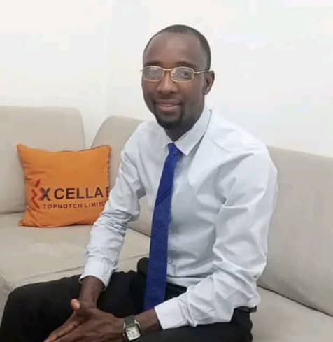 Stanley Ugagbe Man in a light blue shirt and blue tie sits on a beige sofa in an office lounge, with an orange cushion in the background.