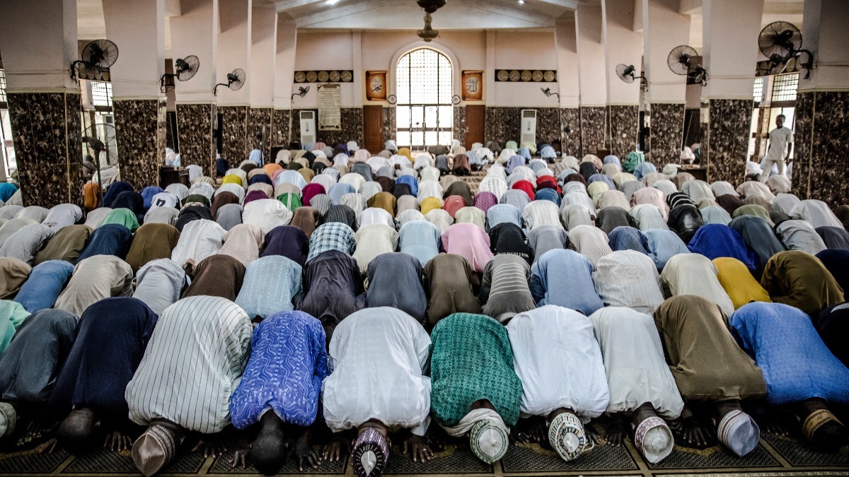 muslim men praying - THEWILL NEWS MEDIA Congregation of Muslims praying in a mosque, bowing in unison during prayer in a crowded prayer hall with tiled walls and arched window at the front, seen from behind.