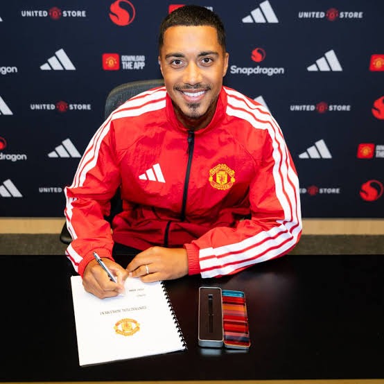 Man wearing a red Manchester United jacket sits at a desk, signing a document with a pen; a notebook and a smartphone lie on the table in front of him.