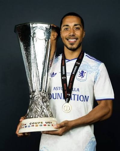 Athlete in a white and light-blue jersey smiles while holding a tall silver cup trophy; a medal hangs around his neck.