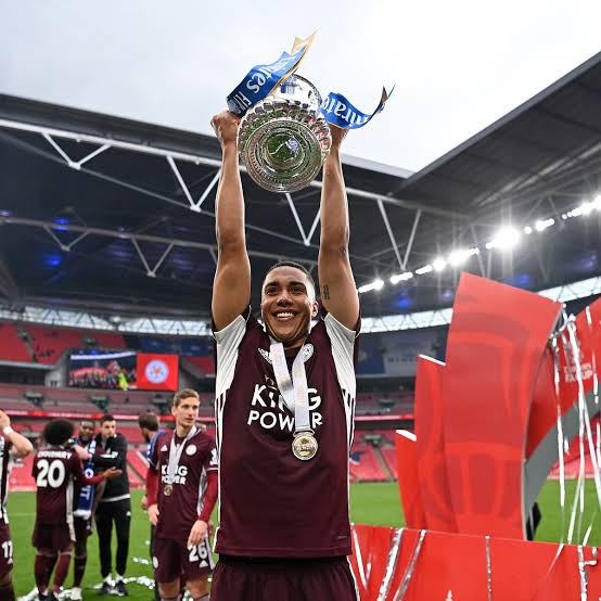 Soccer player in maroon jersey raises a trophy above his head in a stadium as teammates celebrate in the background.