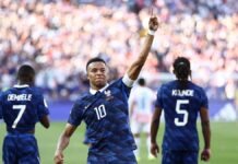 Mbappe Fires France Into World Cup Quarter-Finals After Narrow Win Over Paraguay Soccer player in blue with number 10 raises his right arm in celebration on the field, flanked by teammates wearing numbers 7 and 5.