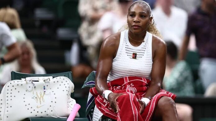 Serena Williams Serena Williams sits courtside on a bench, wearing a white sleeveless top and holding a red towel, with a white studded bag beside her.