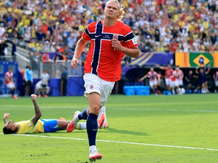 Haaland Norwegian soccer player in red jersey runs on the field while a teammate lies on the grass and spectators cheer in the stands Behind them, a Brazilian flag banner is visible.