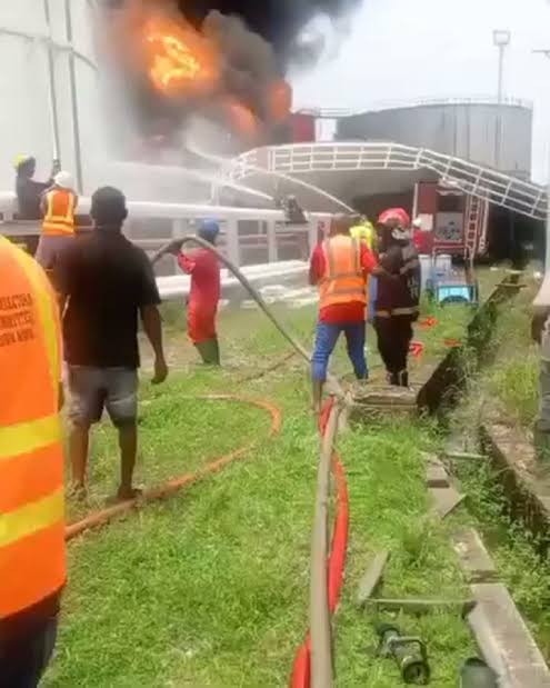 Firefighters in orange vests battle a warehouse blaze, spraying water as thick black smoke rises from the building.