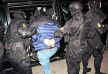 Morocco Foils Major Daesh Terror Plot, Arrests 10 Suspects Across Seven Cities Suspect in a blue jacket is handcuffed and escorted by four masked tactical police officers into a black SUV.