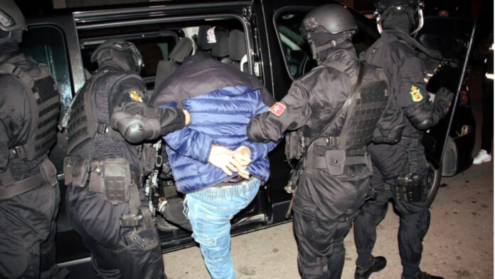 Morocco Foils Major Daesh Terror Plot, Arrests 10 Suspects Across Seven Cities Suspect in a blue jacket is handcuffed and escorted by four masked tactical police officers into a black SUV.