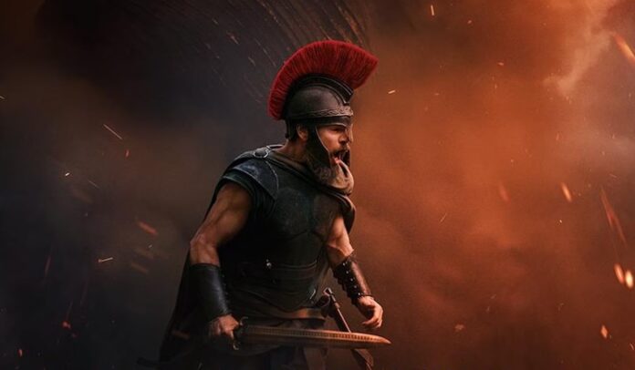 Christopher Nolan’s “The Odyssey” Roman warrior in black armor with a red-plumed helmet readies a sword amid fiery smoke and sparks on a battlefield scene