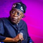 Tinubu Orders EFCC to Unfreeze Osun Account Male performer on stage, wearing a dark pinstripe outfit and a head wrap, holding a microphone under purple lighting