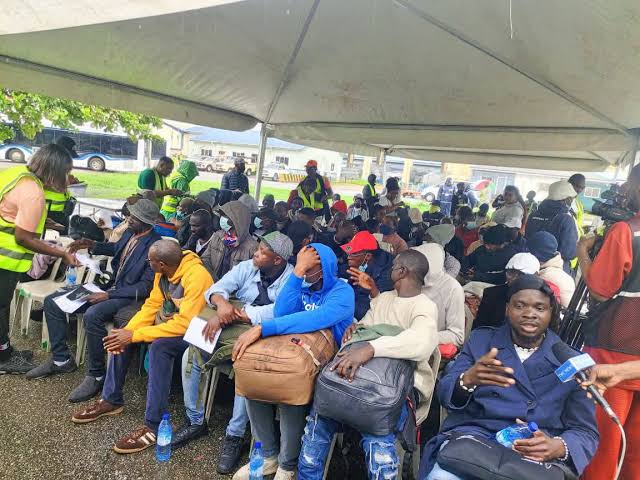 South Africa Returnees People seated under a large canopy at an outdoor aid event; volunteers in reflective vests distribute papers and supplies.