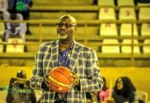 NBBF President Kida Rallies Nigerians To Back D’Tigers Ahead Of World Cup Qualifiers Man in a blue plaid blazer holding a basketball on an indoor court with yellow bleachers behind him.