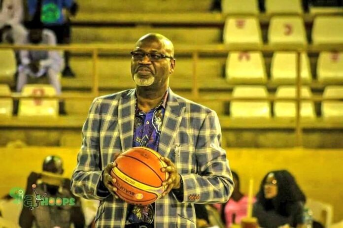 Musa Kida Man in a blue plaid blazer holding a basketball on an indoor court with yellow bleachers behind him.