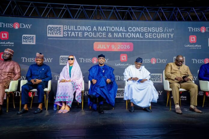 Seven people in traditional attire sit on a stage in front of a large backdrop reading 'Building a National Consensus for State Police & National Security' and displaying logos; panel appears to be at a media briefing or conference.