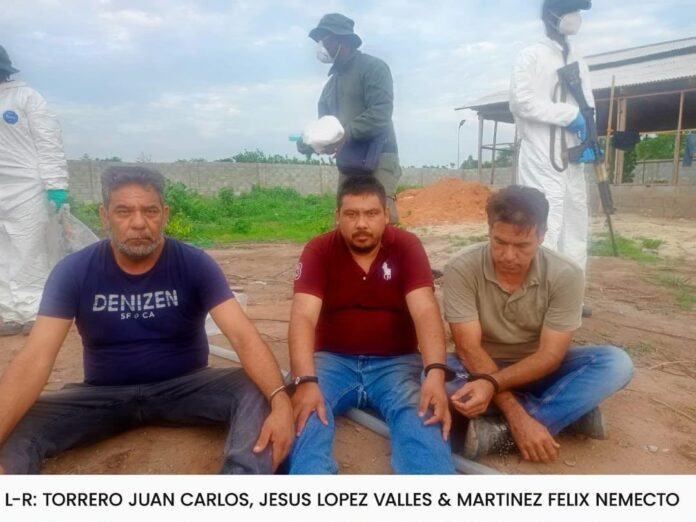Three men sit on a dirt ground with hands resting on their knees, while people in white hazmat suits stand in the background.