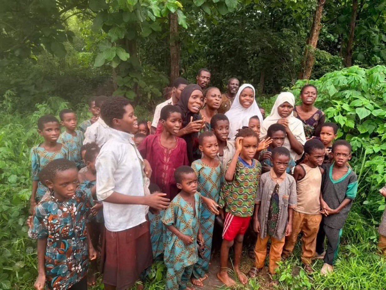 Group of African children and adults gathered outdoors among green trees, looking toward the camera for a group photo.