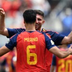 Merino’s Late Strike Sends Spain Past Belgium Into World Cup Semi-Finals Soccer players celebrate a goal with a hug; the back of player Merino wearing number 6 is visible on a red striped jersey.
