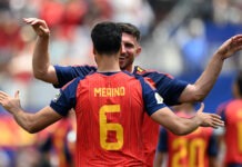 Merino’s Late Strike Sends Spain Past Belgium Into World Cup Semi-Finals Soccer players celebrate a goal with a hug; the back of player Merino wearing number 6 is visible on a red striped jersey.