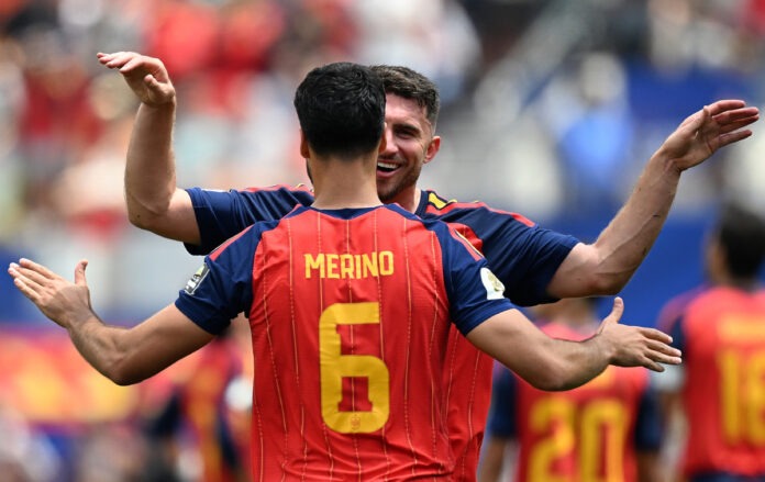 Soccer players celebrate a goal with a hug; the back of player Merino wearing number 6 is visible on a red striped jersey.