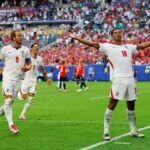 Bellingham Double Fires England Past Norway Into World Cup Semi-Finals England soccer players celebrate a goal on a green pitch with outstretched arms as fans cheer in the stadium stands behind them.